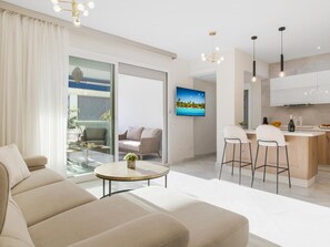 A modern living room with a sofa, a glass table, and a TV. There is a kitchen area with bar stools and a bottle of champagne. - 3 Dormitorios en Playas del Duque Ref C2 111 (Marbella)
