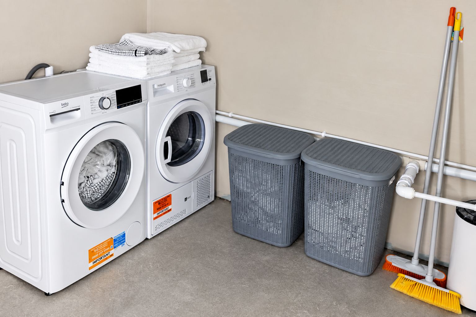 Un espace buanderie comprenant deux machines à laver, deux paniers à linge gris et un balai.