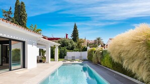 Uma piscina moderna com superfície de água azul cristalina, rodeada por um deck pavimentado e um edifício branco com grandes portas de vidro.