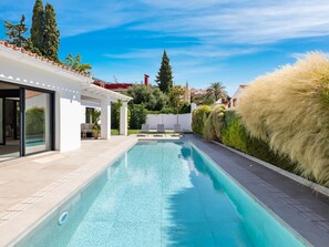 A modern swimming pool with a clear blue water surface, surrounded by a paved deck and a white building with large glass doors. - Villa LAS Palmeras (Marbella)