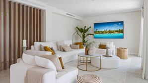 Uma sala de estar moderna com um sofá branco, uma mesa de centro redonda e uma pintura grande de uma praia tropical.