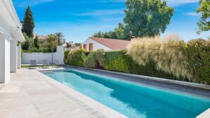 Piscina com deck ladrilhado, rodeada por vegetação e um edifício branco.