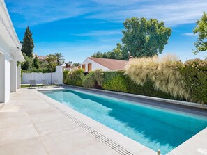 A swimming pool with a tiled deck, surrounded by greenery and a white building. - Villa LAS Palmeras (Marbella)
