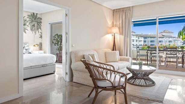 Ein Hotelzimmer mit Balkon, einem Bett, einer Couch, einem Korbstuhl und einem runden Tisch.
