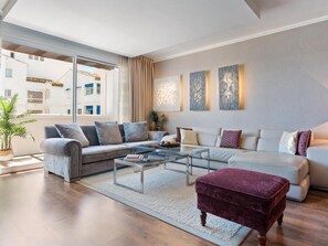 A modern living room with a grey sofa, a coffee table, and a red ottoman. There are abstract wall art pieces and a large window providing a view of buildings outside. - 03 Dormitorios ref Casa Granada 146 (Marbella)