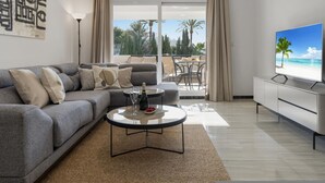 Uma sala de estar moderna com um sofá cinzento, uma mesa de centro com uma garrafa e copos, uma televisão de tela plana e uma varanda com vista para o mar.