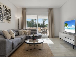 Ein modernes Wohnzimmer mit grauem Sofa, einem Couchtisch mit Flasche und Gläsern, einem Flachbildfernseher und einem Balkon mit Meerblick.