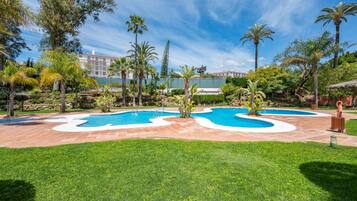 Zona de piscina ajardinada con palmeras y un edificio al fondo.
