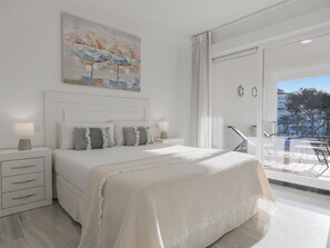 Ein modernes Schlafzimmer mit einem großen Bett, einem Nachttisch, einer kleinen Kommode und Blick ins Freie durch eine Schiebetür.