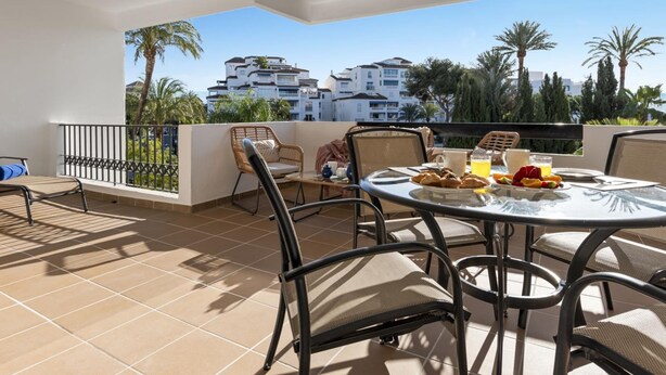 Terraço com mesa posta para o café da manhã, com vista para uma área residencial com palmeiras e edifícios.