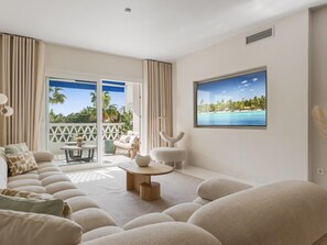 Ein modernes Wohnzimmer mit großem Flachbildfernseher, einer Couch, einem Couchtisch und einem Balkon mit Blick auf Palmen.