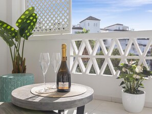 Ein runder Tisch mit einer Weinflasche, zwei Gläsern und einer gewebten Tischmatte auf einem Balkon mit weißem Gitter und Blumentöpfen.