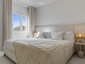 Ein modernes Schlafzimmer mit einem großen Bett, einem Nachttisch mit Lampe und Blick ins Freie durch ein Fenster.