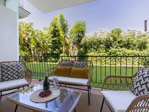 An outdoor seating area with wicker furniture, a glass table, and a view of a well-maintained garden and trees. - Cerca de la Playa ref Adm409 (Marbella)