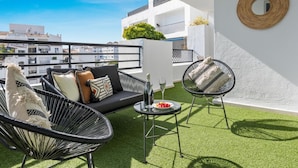 Una terrazza sul tetto con un divano nero, due poltrone in vimini, un tavolino con un bicchiere di vino e uno specchio rotondo appeso al muro.
