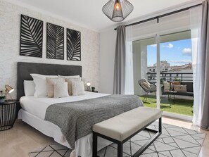 A bedroom with a bed, bedside tables, a bench, and a view of a balcony with outdoor furniture. - Confortable y c Ntrico en Terrazas de Ban s (Marbella)