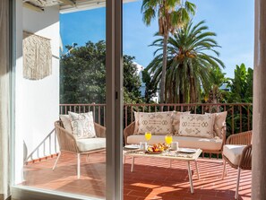 Ein Balkon mit Korbmöbeln, einem Tisch mit Obst und Getränken und Blick auf Palmen und ein Gebäude.