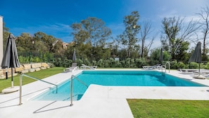 Ein rechteckiger Pool mit Metall-Sicherheitsgeländer, umgeben von einer gepflasterten Liegefläche. Liegest ühle und Sonnenschirme befinden sich rund um den Pool.