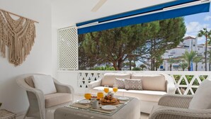Un espace extérieur ensoleillé, aménagé avec des meubles en osier, une table en verre sur laquelle sont disposés des aliments pour le petit-déjeuner, et offrant une vue sur les arbres et les bâtiments.