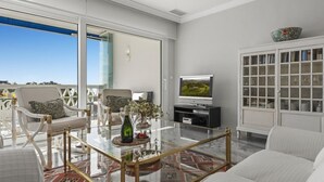 A modern living room with a glass coffee table, white sofas, a television, and a view of the outdoors. - Vista mar y Piscina en Puerto Ban s (Marbella)