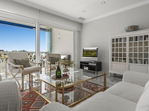 Ein modernes Wohnzimmer mit einem Glastisch, weißen Sofas, einem Fernseher und Blick ins Freie.