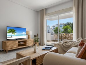 A modern living room with a television displaying a beach scene, a wooden coffee table, a sofa, and a balcony with outdoor furniture. - Apartamento de 2 Dormitorios con Terraza ref 211 (Marbella)