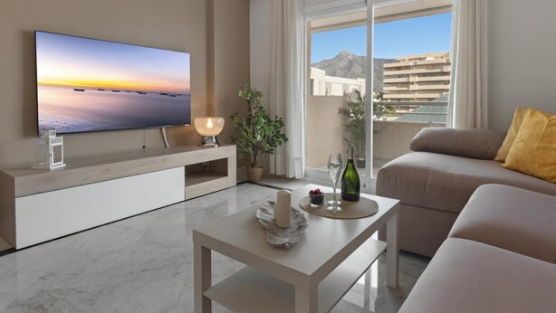Ein modernes Wohnzimmer mit Fernseher, Couchtisch, Sofa und einem Balkon mit Blick auf Gebäude und Berge.