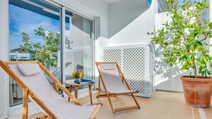 Een balkon met twee houten lounge fauteuils, een tafeltje met een boek en een glas sinaasappelsap, en een bloempot met plant.