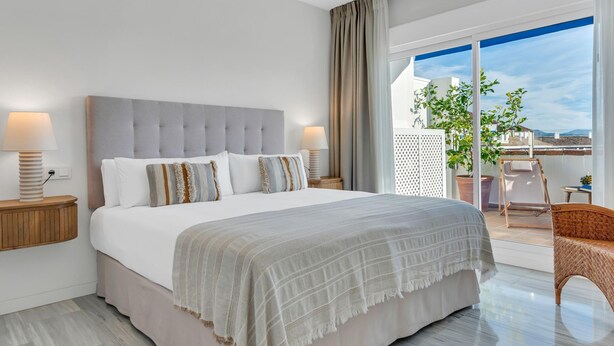 Een moderne hotelkamer met een groot bed, twee nachtkastjes met lampen, een balkon met een rieten stoel en uitzicht op de stad.