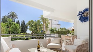 A balcony with a white sofa, two chairs, a small table with a bottle and glasses, and a view of palm trees and buildings. - Medina Garden 201 in Puerto Ban s - Marbella (Marbella)