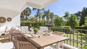 A balcony with a wooden table set for dining, wicker chairs, and a view of palm trees and buildings. - Medina Garden 201 in Puerto Ban s - Marbella (Marbella)