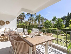 A balcony with a wooden table set for dining, wicker chairs, and a view of palm trees and buildings. - Medina Garden 201 in Puerto Ban s - Marbella (Marbella)