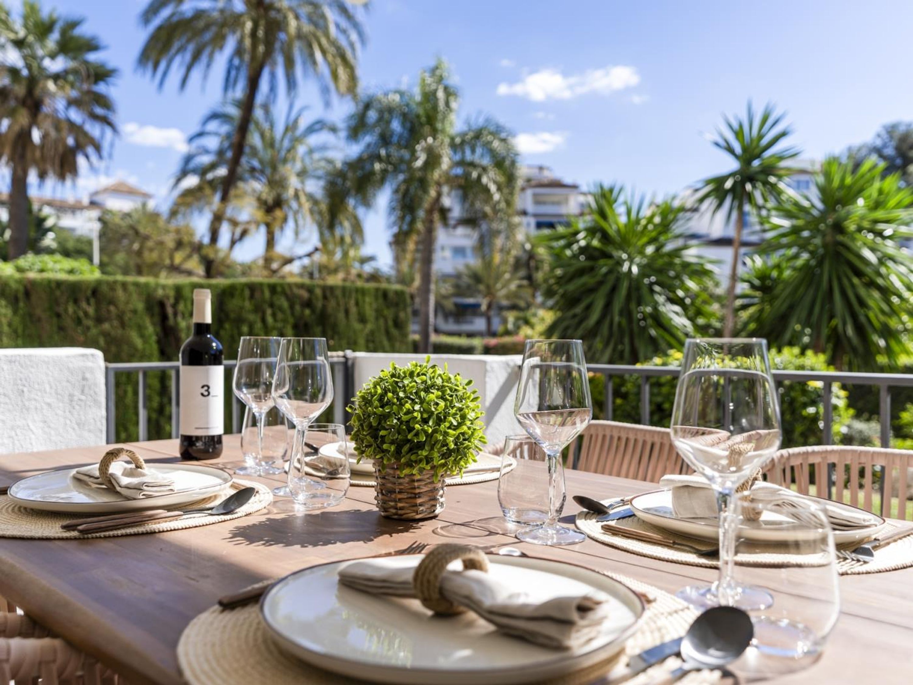 Mesa de comedor al aire libre con platos, vasos y una botella, rodeada de exuberante follaje y palmeras.