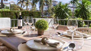 An outdoor dining table set with plates, glasses, and a bottle, surrounded by lush greenery and palm trees. - Medina Garden 201 in Puerto Ban s - Marbella (Marbella)