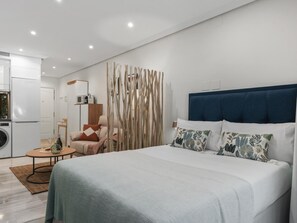 Ein modernes Schlafzimmer mit Bett, einem Sessel, einem kleinen Tisch und einer Küchenzeile im Hintergrund.