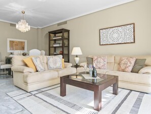 Ein Wohnzimmer mit einem beigen Sofa, einem Glastisch, einer Kandelaber, einem Bücherregal und einem Wandkunstwerk.
