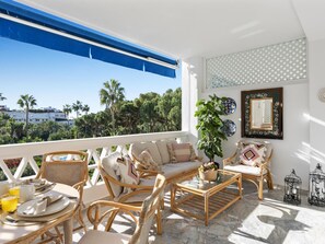 Ein Balkon mit Korbmöbeln, einem Glastisch und Blick auf Palmen und Gebäude.