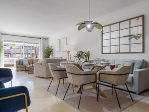 Ein modernes Wohnzimmer mit einem Esstisch mit Glasplatte, beigen Sofas und einem großen Fenster mit Blick auf Gebäude.