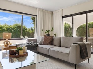 A modern living room with a sofa, a glass coffee table, and a view of greenery outside. - Villa Byrons in Marbella (Marbella)