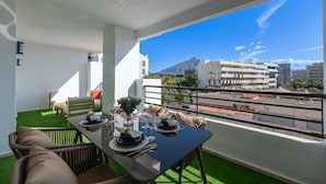 Een balkon met een eettafel gedekt voor twee personen, een bank en uitzicht op een berg en gebouwen.