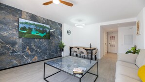 A modern living room with a flat-screen TV mounted on a marble wall, a glass coffee table, a dining area with chairs, and a sofa. - Los Angeles Beach Front Villa (Marbella)