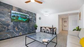 Ein modernes Wohnzimmer mit einem Flachbildfernseher an einer Marmorwand, einem Glastisch, einem Essbereich mit Stühlen und einem Sofa.