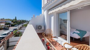 Ein Balkon mit Blick auf eine Straße, ein Korbsessel, ein Bett mit einem grünen Handtuch und eine Klimaanlage.