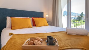 Ein Hotelzimmer mit einem Bett, Kissen, einem Tablett mit Gebäck und Kaffeetassen sowie Blick auf Berge durch das Fenster.