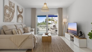 A modern living room with a sofa, coffee table, and TV, a balcony with outdoor furniture, and artwork on the walls. - Beach Puerto Ban s 3 Br Apartment (Marbella)