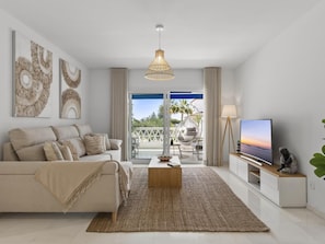 A modern living room with a sofa, coffee table, and TV, a balcony with outdoor furniture, and artwork on the walls. - Beach Puerto Ban s 3 Br Apartment (Marbella)