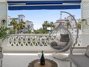 A balcony with a hanging chair, a bottle of champagne, and a view of buildings and palm trees. - Beach Puerto Ban s 3 Br Apartment (Marbella)