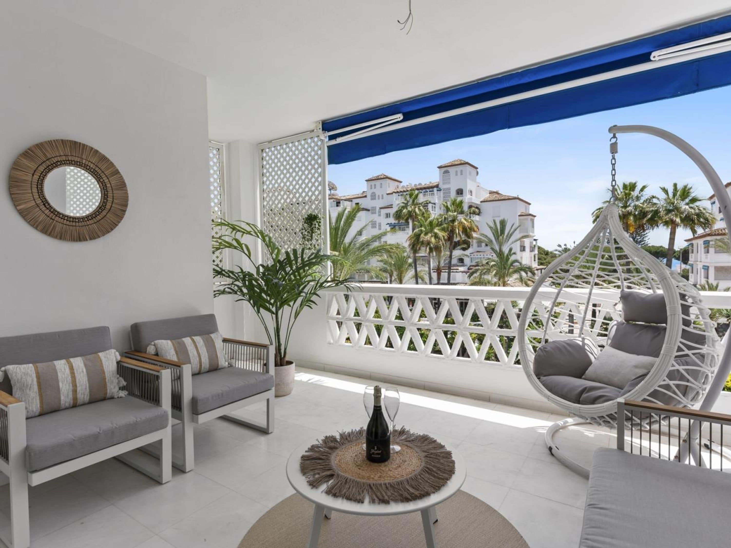 A balcony with a white sofa, a wicker chair, and a table with a bottle and glass.