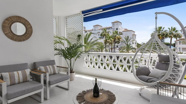 A balcony with a white sofa, a wicker chair, and a table with a bottle and glass. - Beach Puerto Ban s 3 Br Apartment (Marbella)