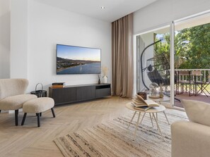 Ein modernes Wohnzimmer mit großem Flachbildfernseher, einer Couch, einem Couchtisch und Blick auf einen Balkon mit Gartenmöbeln.